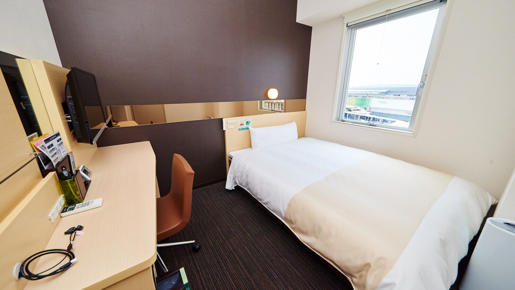 A hotel room with a bed, a desk, a chair, a small table with a bottle and a phone, a wall-mounted TV, and a window with a view of buildings.