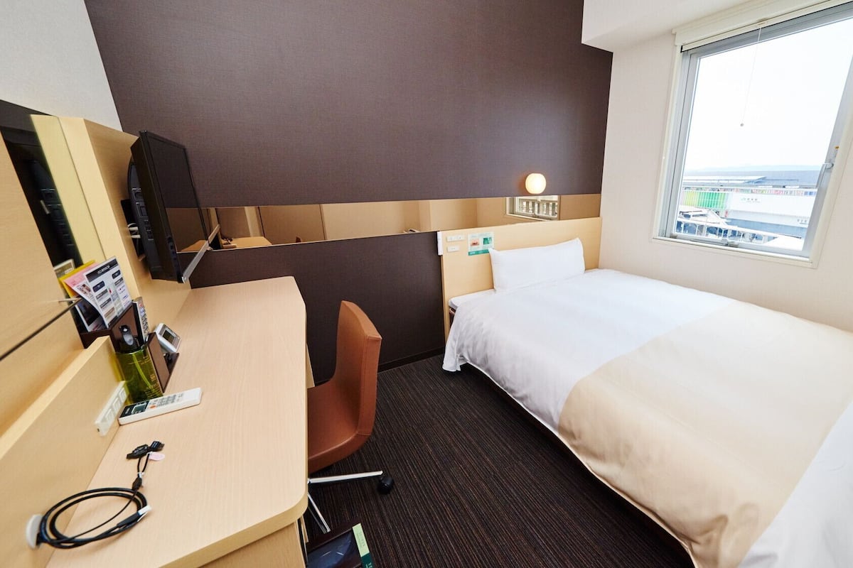 Ein Hotelzimmer mit Bett, Schreibtisch, Stuhl, einem kleinen Tisch mit Flasche und Telefon, einem wandmontierten Fernseher und einem Fenster mit Blick auf Gebäude.