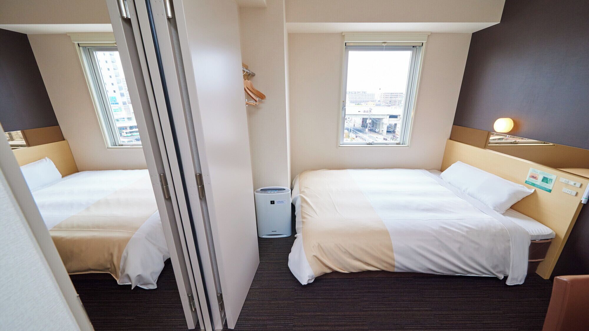 Ein Hotelzimmer mit zwei Betten, einem Fenster mit Stadtblick, einer kleinen Mülleimer und einem Nachttisch mit Lampe.