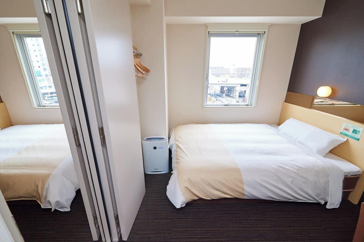 Ein Hotelzimmer mit zwei Betten, einem Fenster mit Stadtblick, einer kleinen Mülleimer und einem Nachttisch mit Lampe.