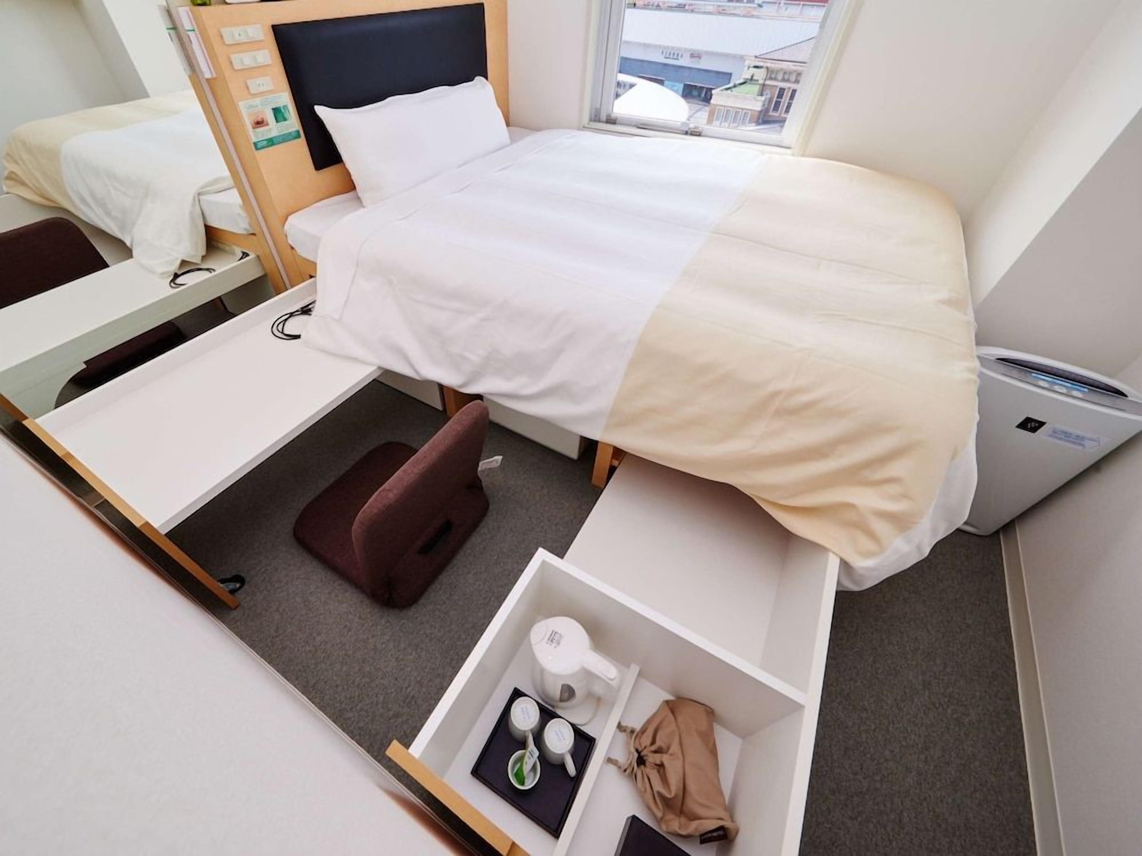 A hotel room with a bed, bedside table, chair, and a small storage unit containing toiletries.