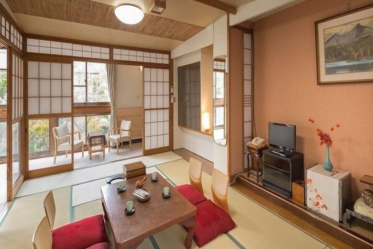 Ein traditioneller japanischer Raum mit Tatami-Boden, einem niedrigen Holztisch mit Kissen, einem Fernseher und einem Bild an der Wand.