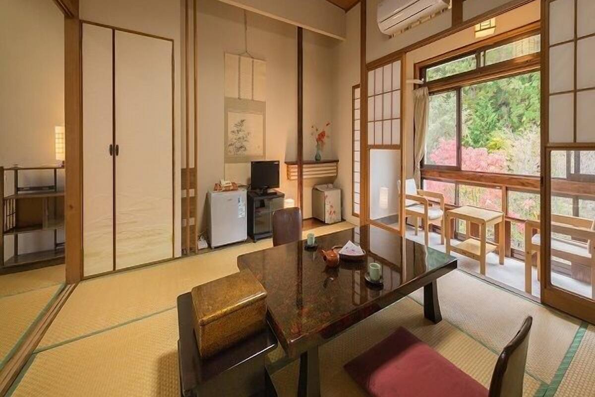 Ein traditioneller japanischer Raum mit Tatami-Böden, einem niedrigen Tisch mit Kissen, einem Fernseher und Blick ins Grüne.