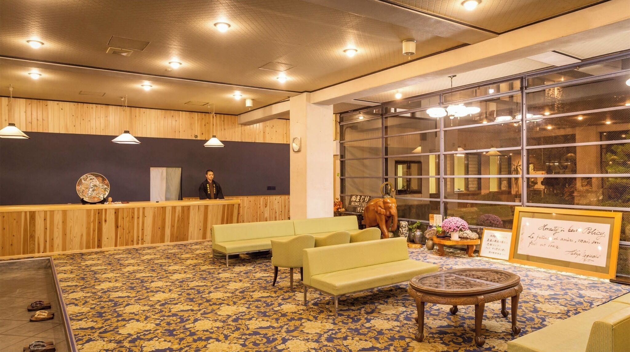 Uma área de recepção com balcão de madeira, um recepcionista e um espaço de estar com sofás e uma mesa de centro.