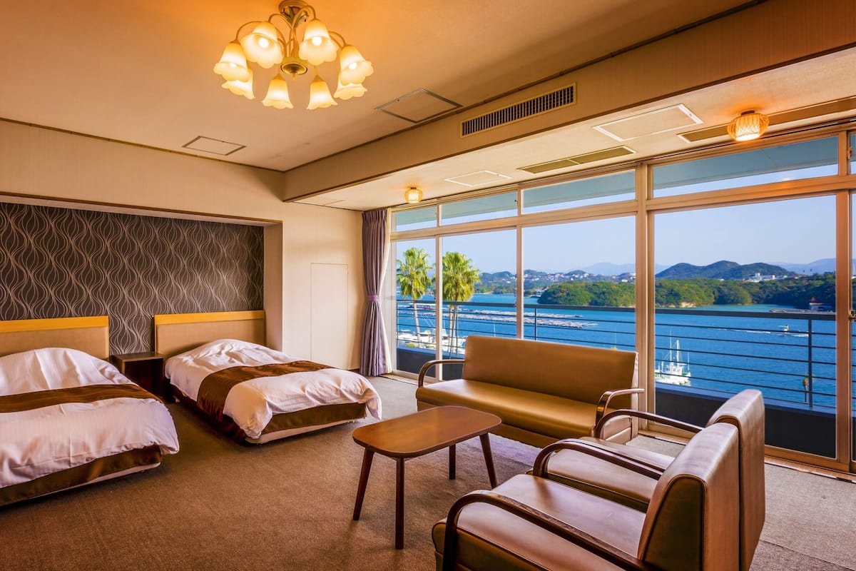 A hotel room with two beds, a sofa, a small table, and large windows offering a view of the sea and hills.