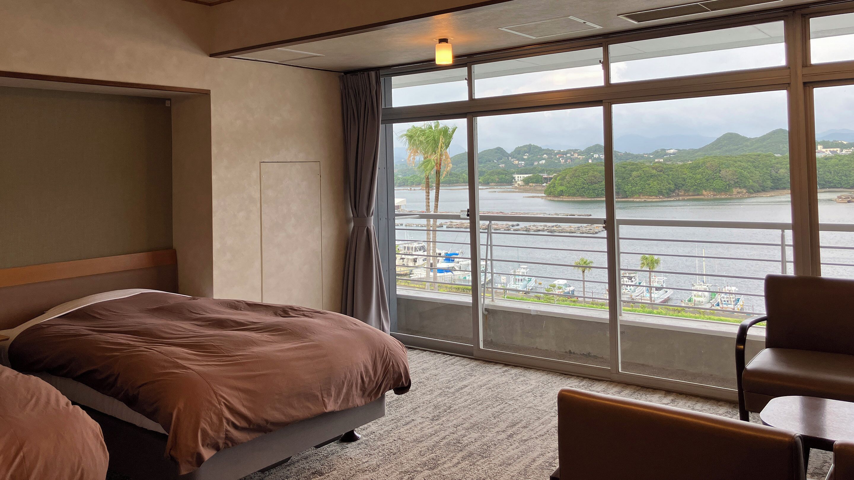 A hotel room with a large window, a bed, a chair, and a view of a river and hills.