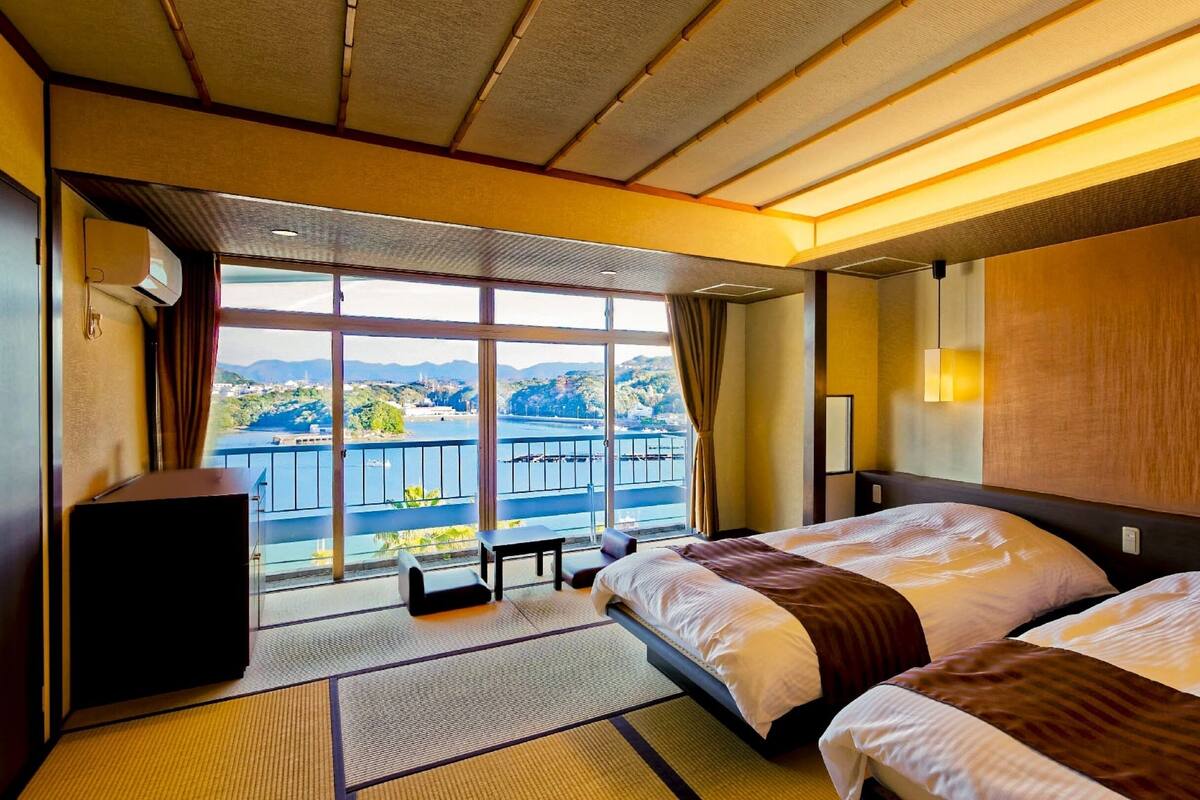 A hotel room with two beds, a large window with a view of a body of water and hills, and a small table with a chair.