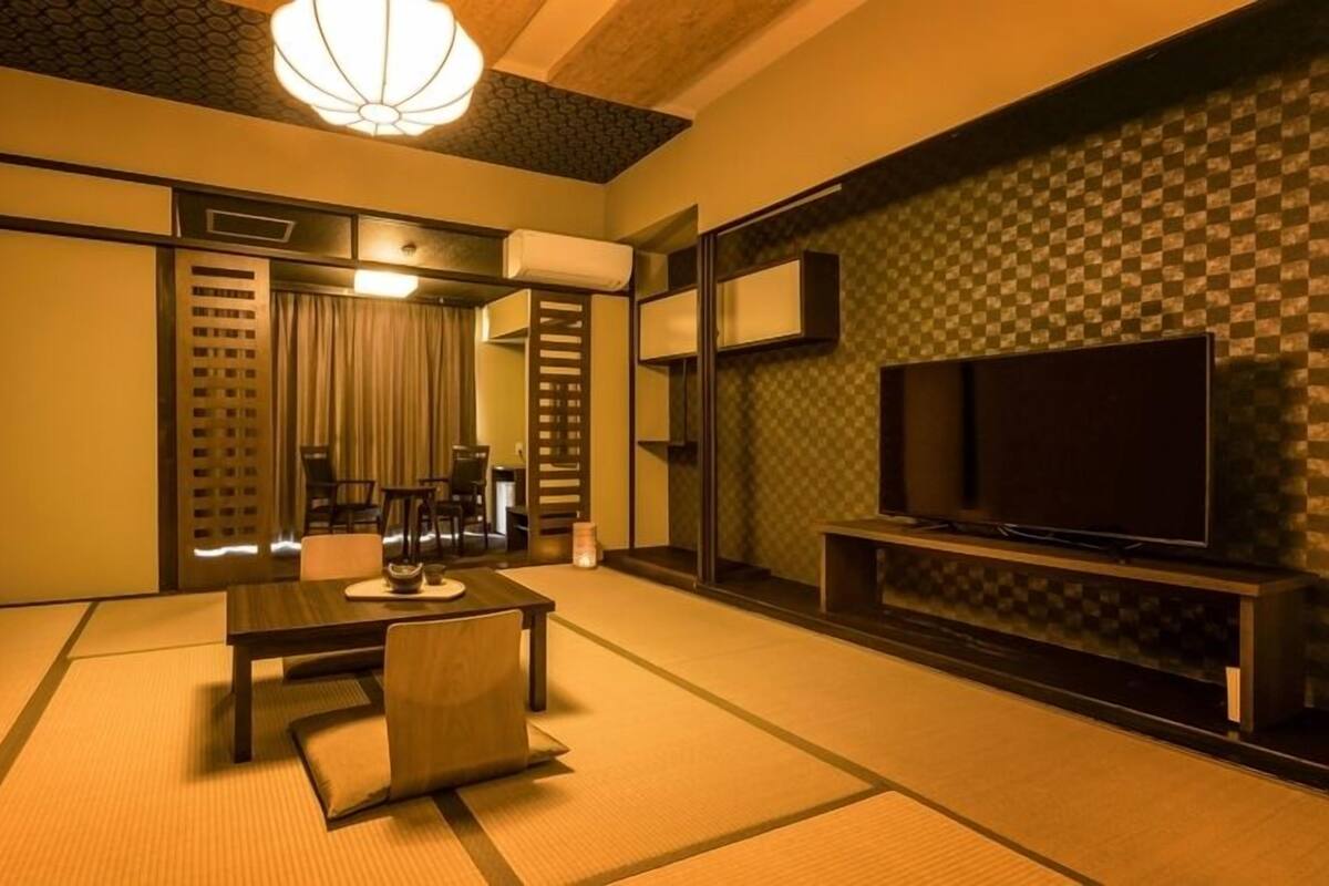 A traditional Japanese-style room with tatami flooring, a low table with cushions, a television on a stand, and a sliding door leading to another room.