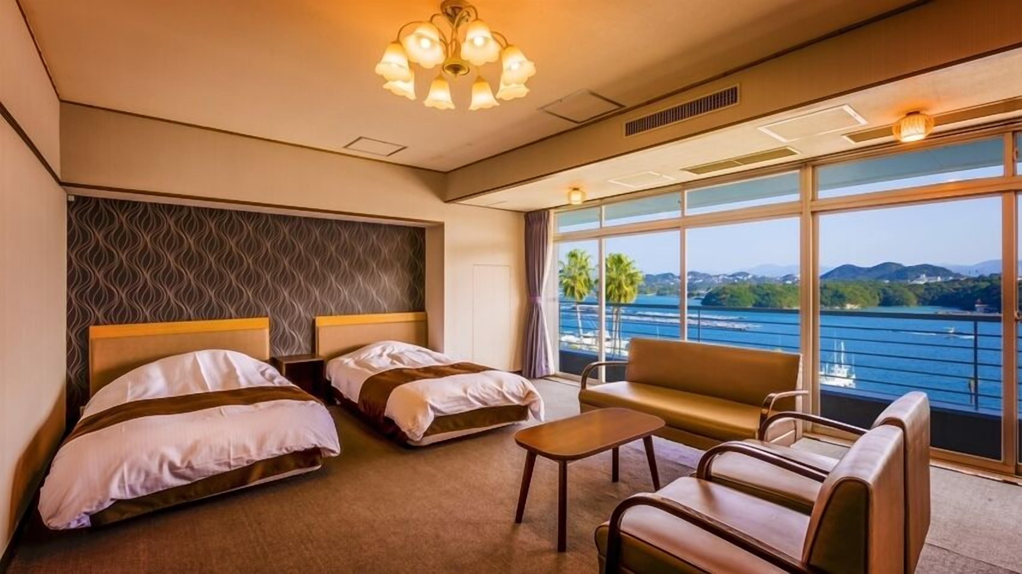 A hotel room with two beds, a sofa, a small table, and large windows with a view of the sea and mountains.