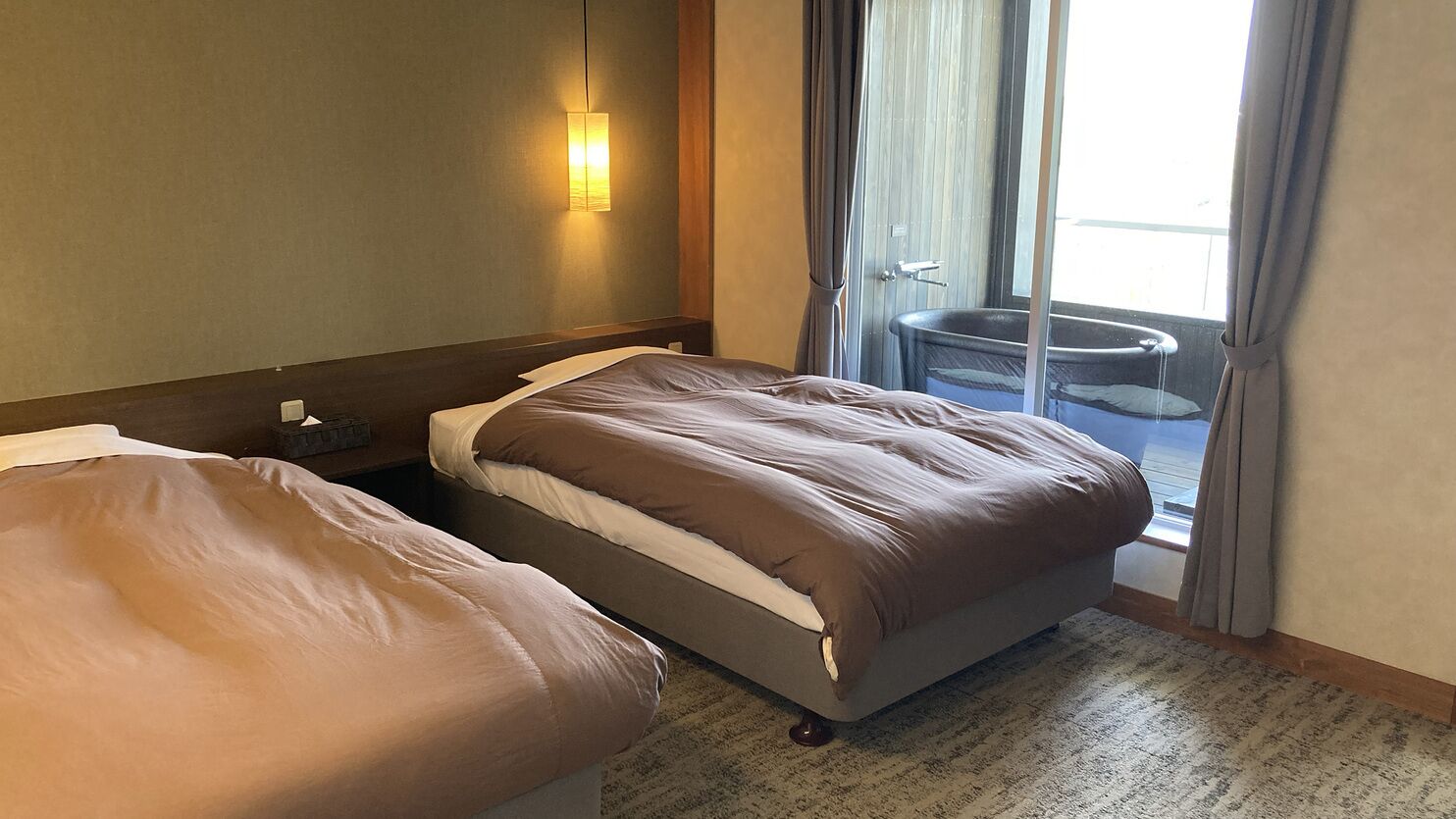 A hotel room with two beds, a nightstand, a wall-mounted lamp, and a view of a pool through a sliding door.