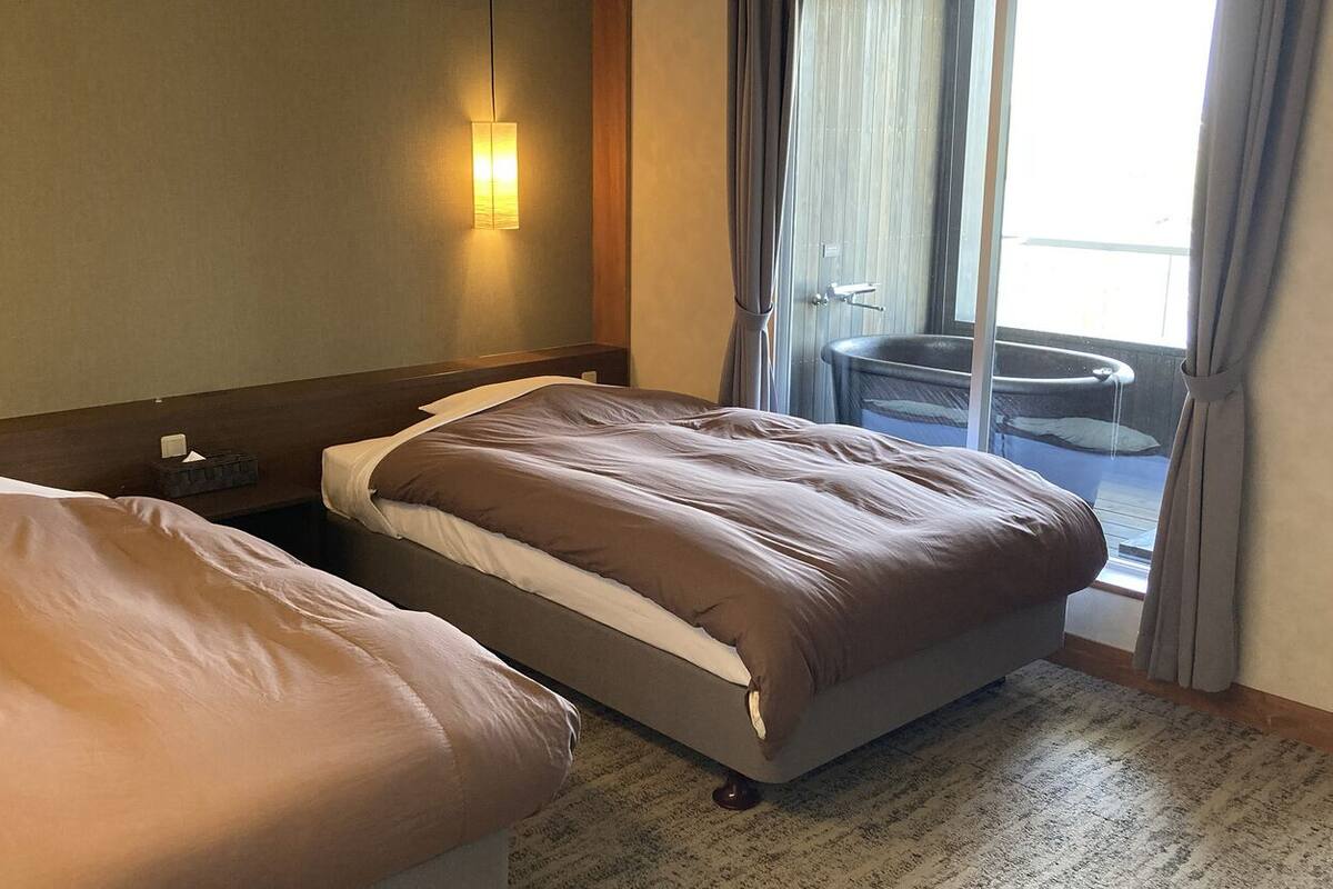 A hotel room with two beds, a nightstand, a wall-mounted lamp, and a view of a pool through a sliding door.