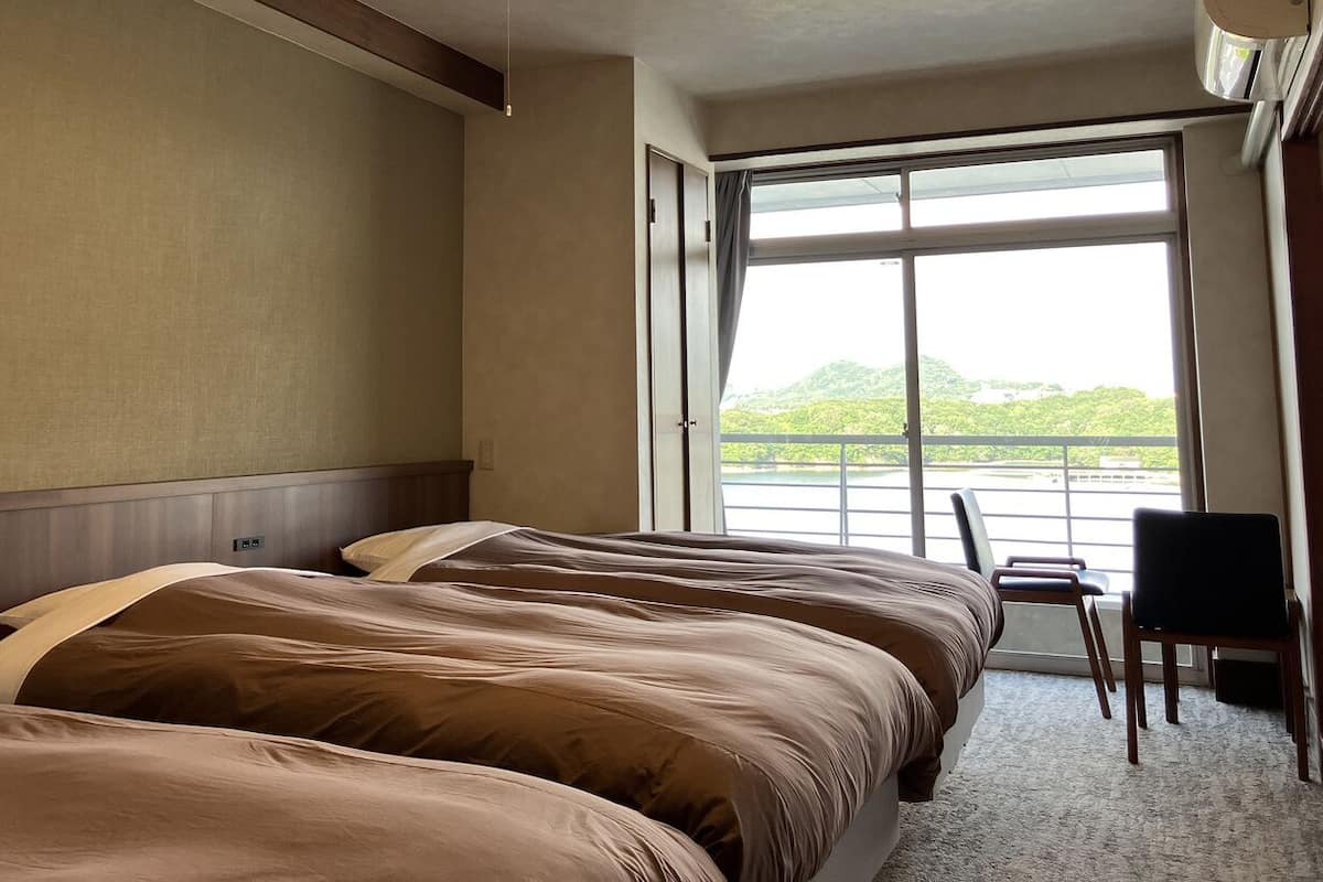A hotel room with two beds, a chair, and a large window offering a view of greenery.