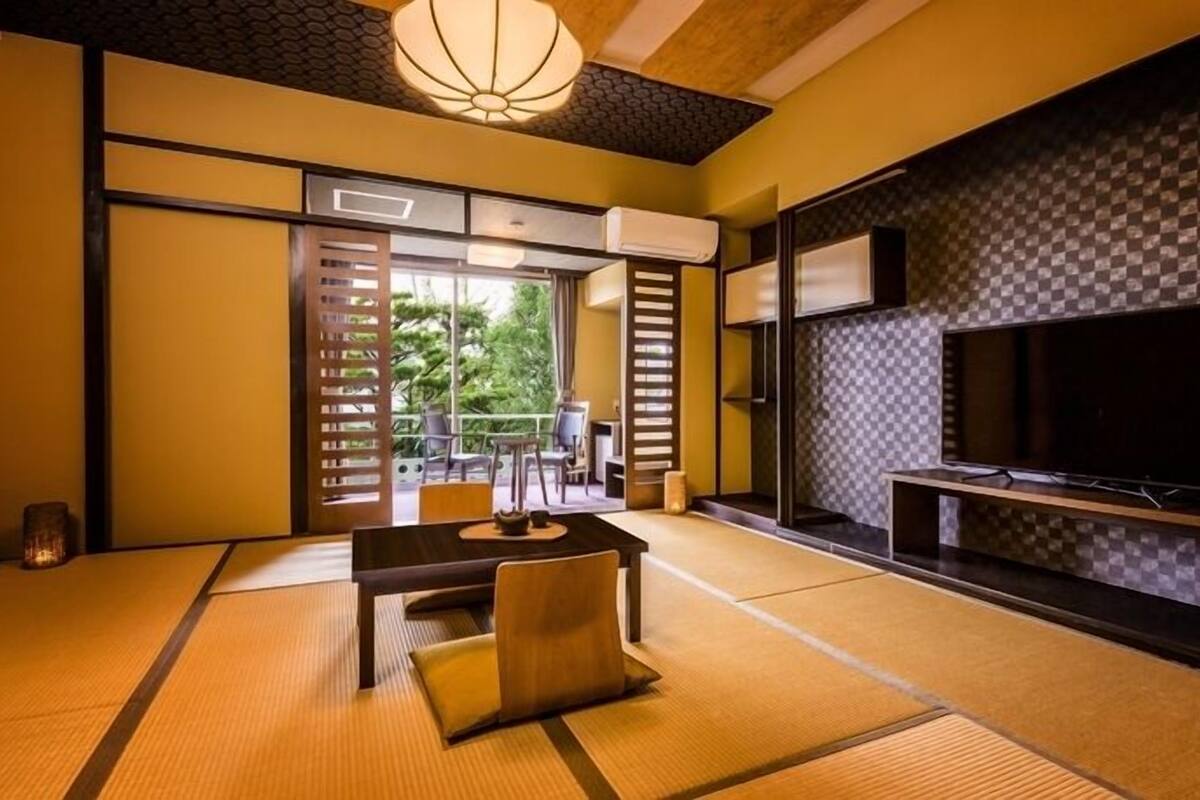 A traditional Japanese room with tatami flooring, a low table with cushions, sliding doors, a television, and a view of greenery outside.