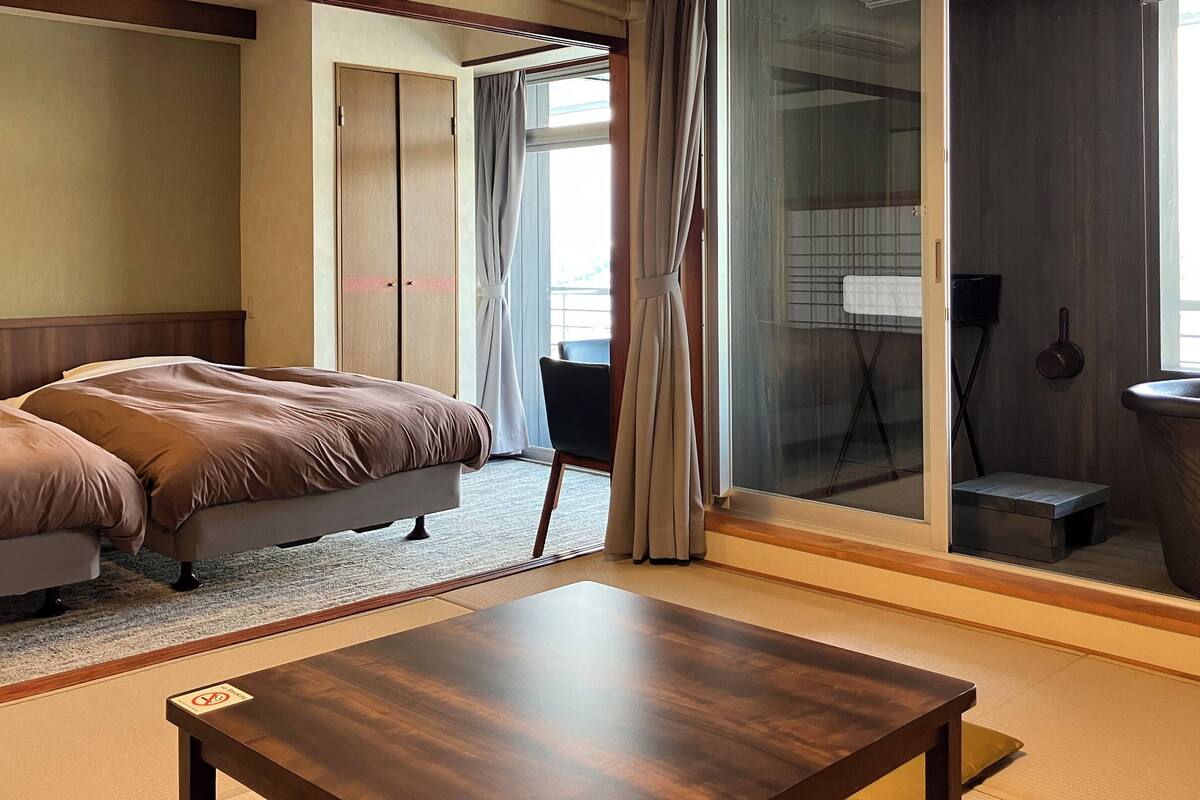 A hotel room with a bed, a small table, a chair, a sliding door, and a view of the outside.