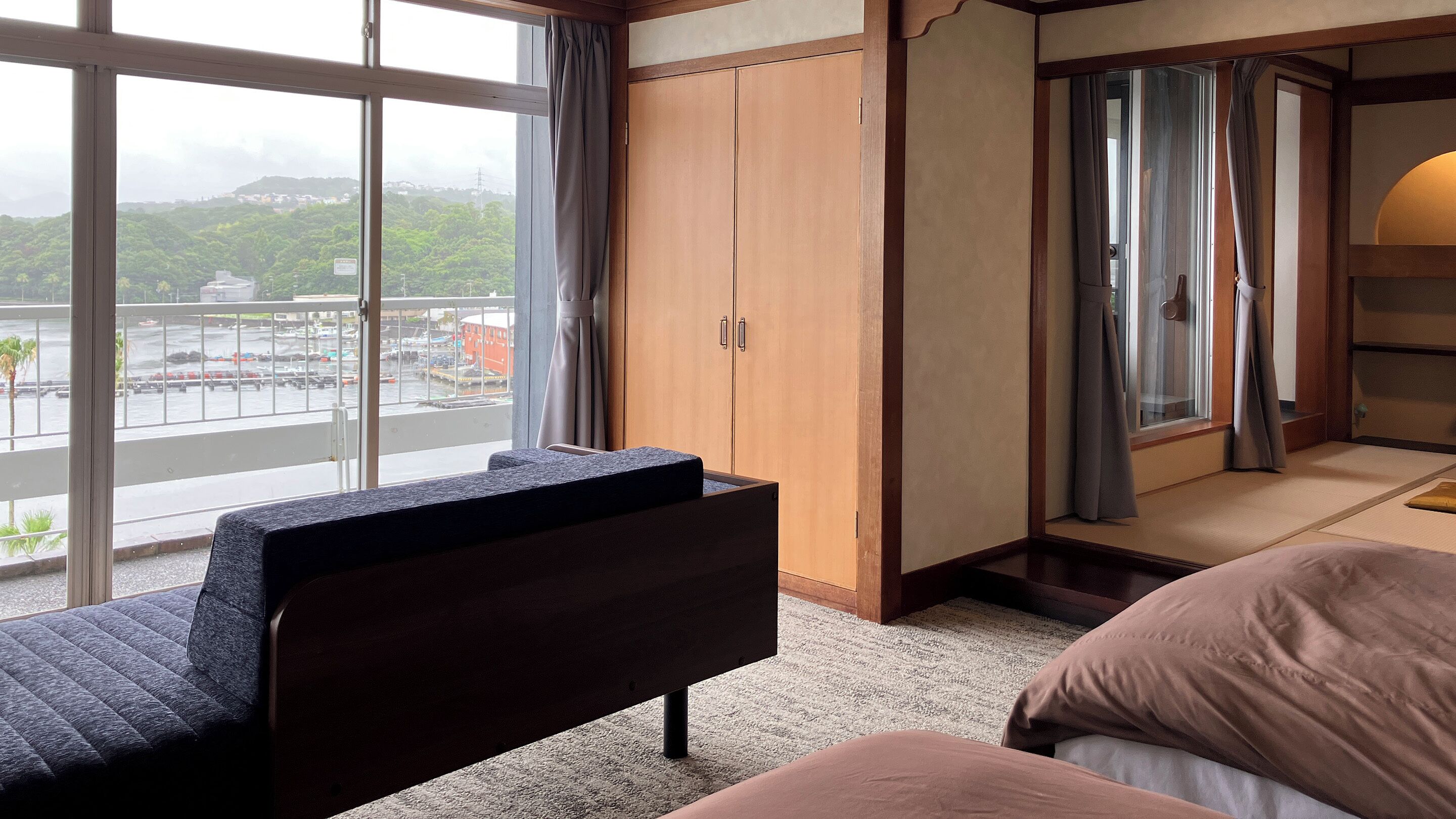 A hotel room with a bed, a sofa, a wardrobe, and a view of a cityscape through large windows.