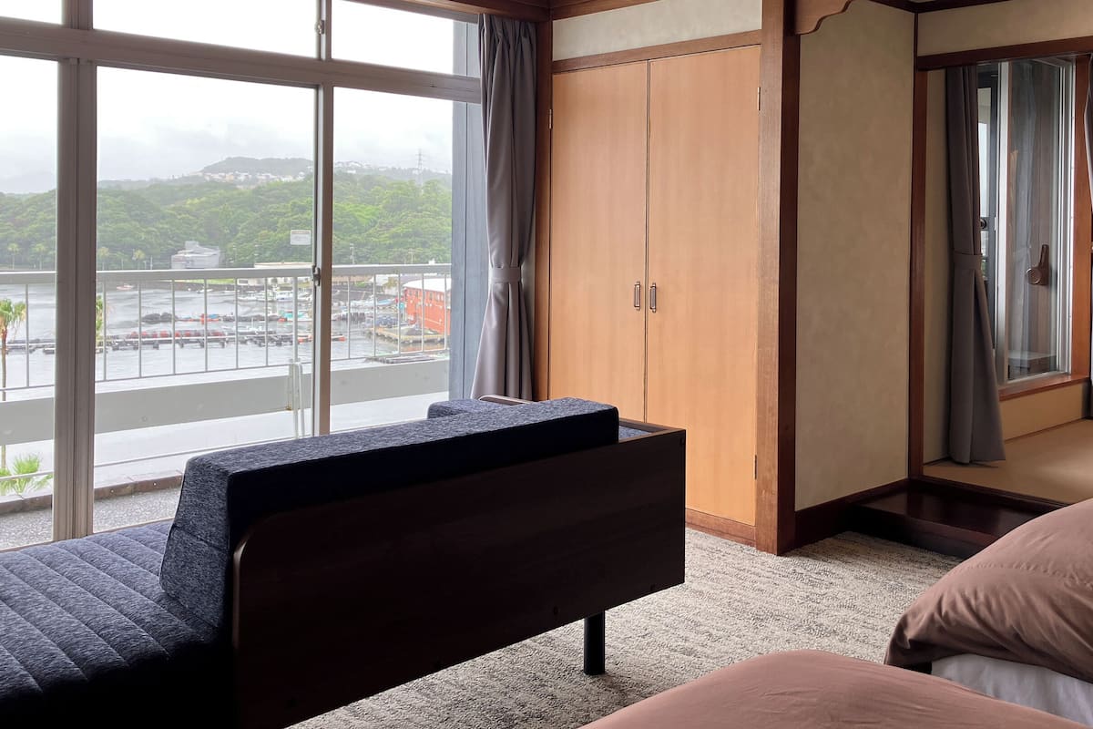 A hotel room with a bed, a sofa, a wardrobe, and a view of a cityscape through large windows.