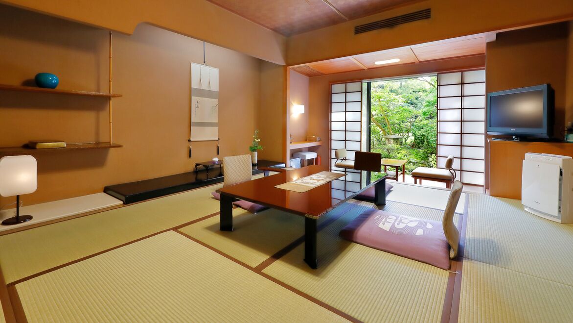 Una habitación japonesa tradicional con suelo de tatami, una mesa baja, sillas, un televisor y vistas al exterior con vegetación.