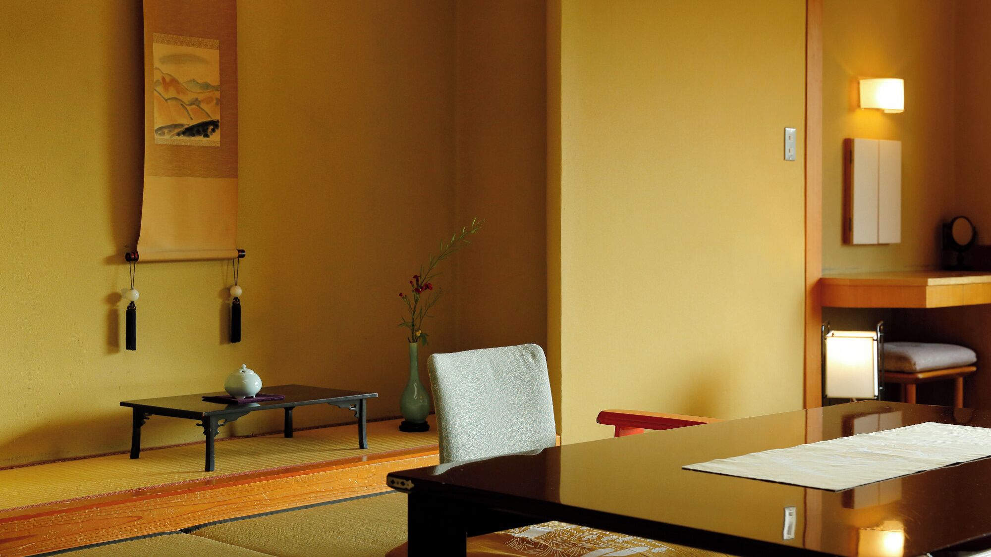 Una habitación de hotel con una mesa de madera, una silla, un escritorio pequeño, un florero con flores y un estante con lámpara.