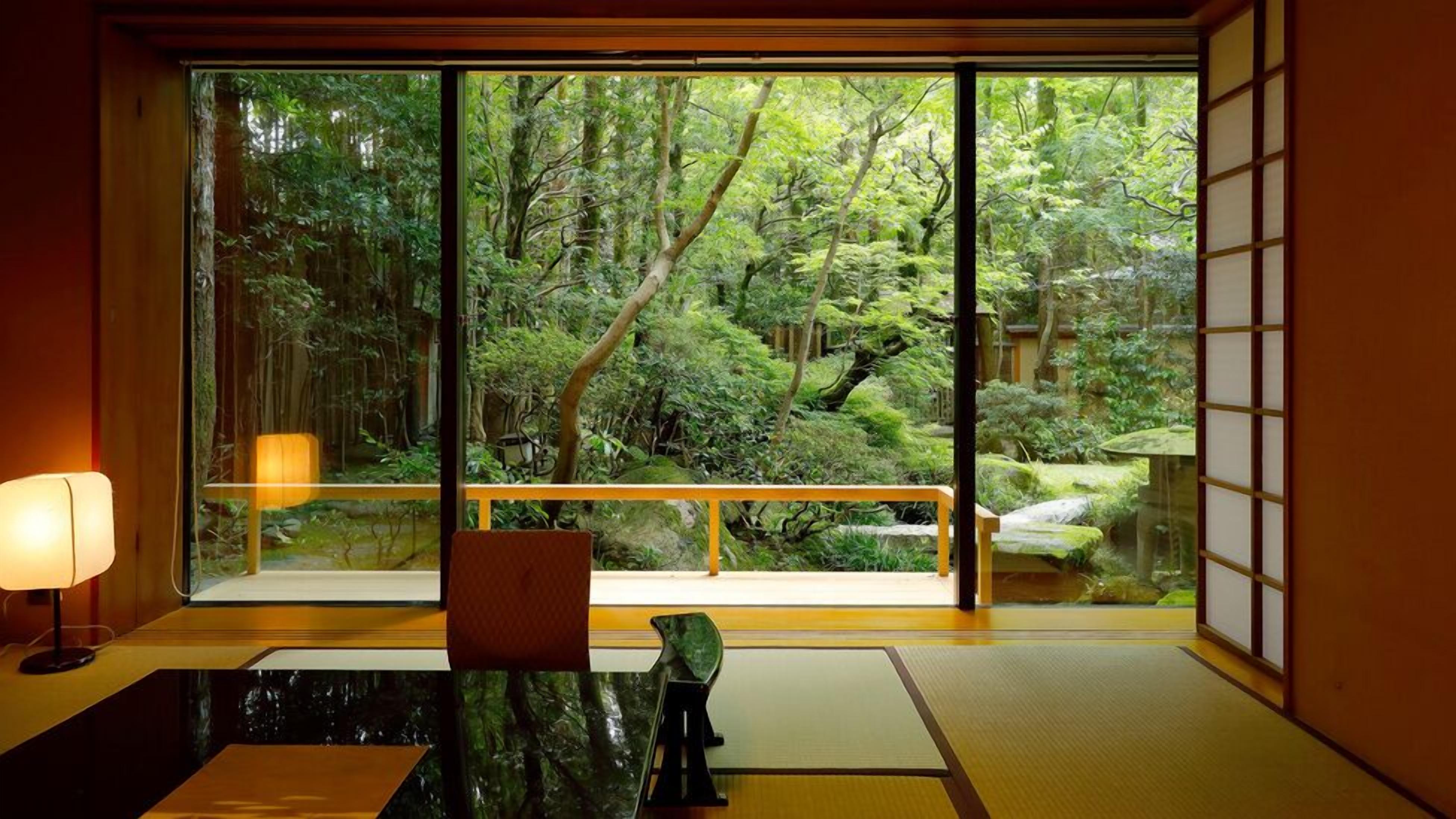 Una habitación japonesa tradicional con suelo de tatami, una mesa baja, un banco y un ventanal con vistas a un jardín arbolado.