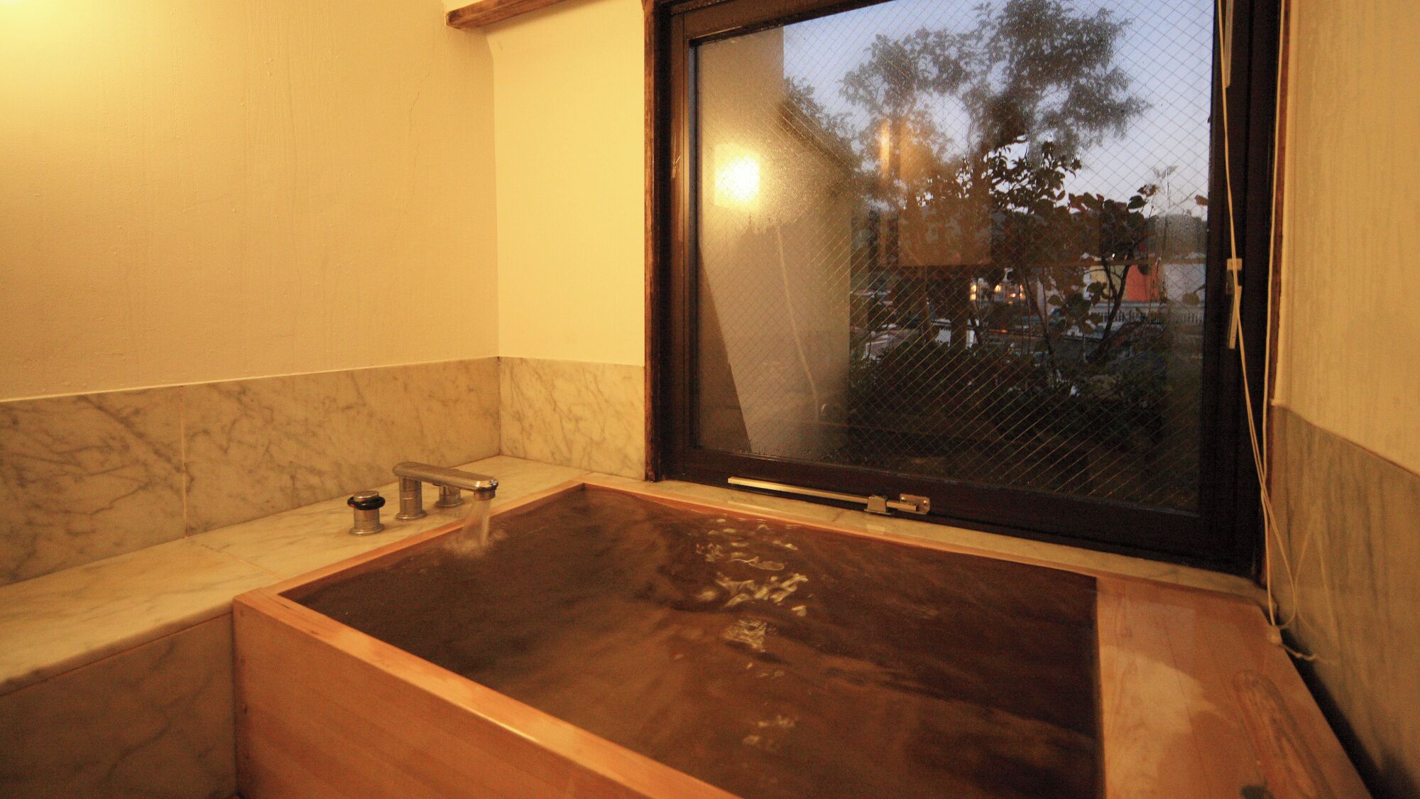 Un baño con una bañera de madera, encimera de mármol y ventana con vistas a árboles y edificios.