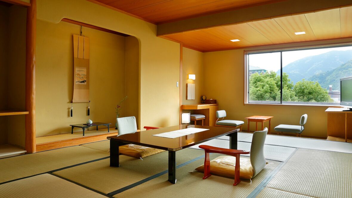 Una habitación japonesa tradicional con suelo de tatami, una mesa baja, sillas, un televisor y una ventana con vistas a zonas verdes.