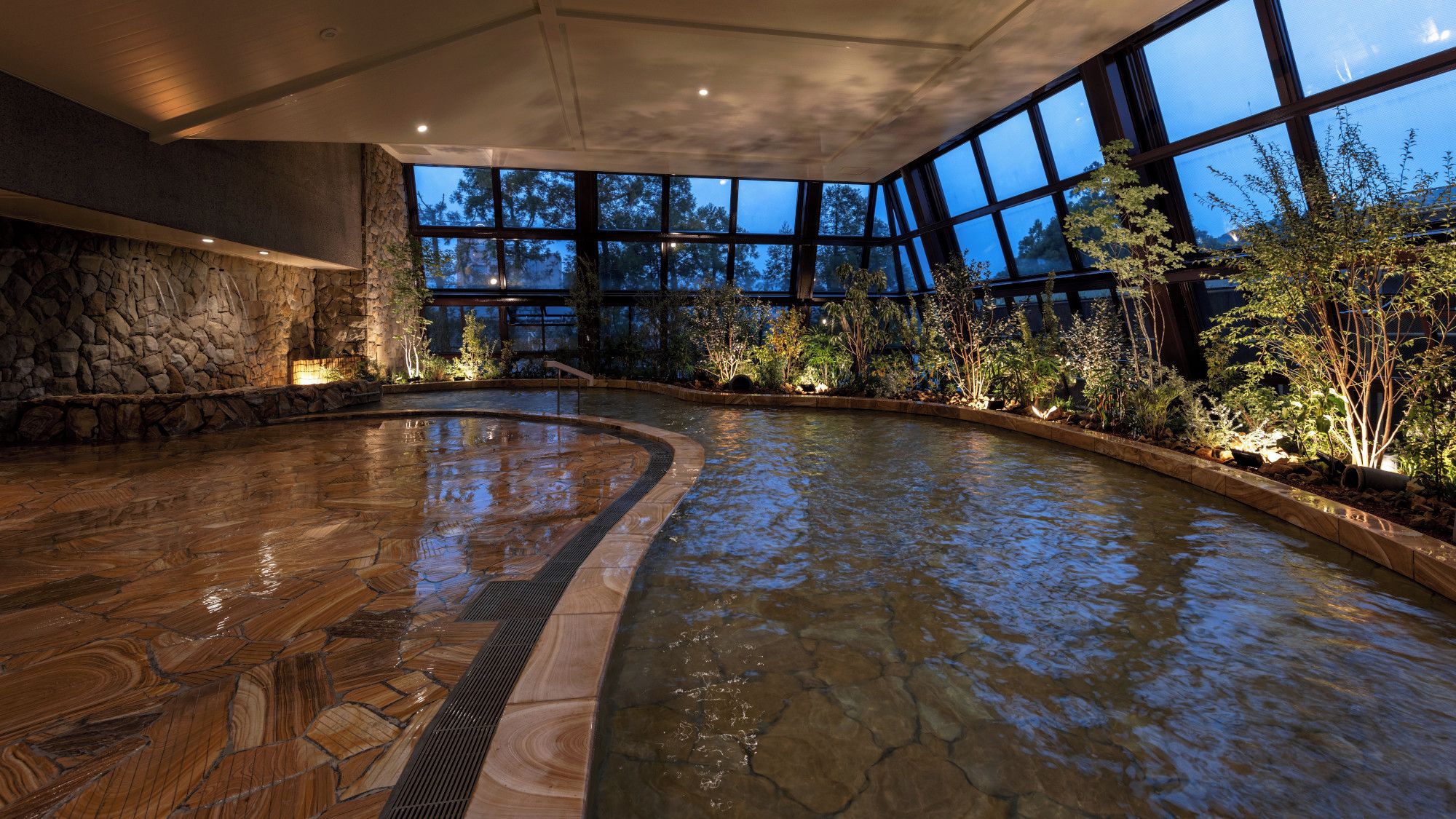 An indoor swimming pool with a stone wall and a view of trees through large windows.