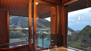 A modern bathroom with a large bathtub, a shower, and a view of a mountainous landscape. - Hotel Zuiyo (Atami)