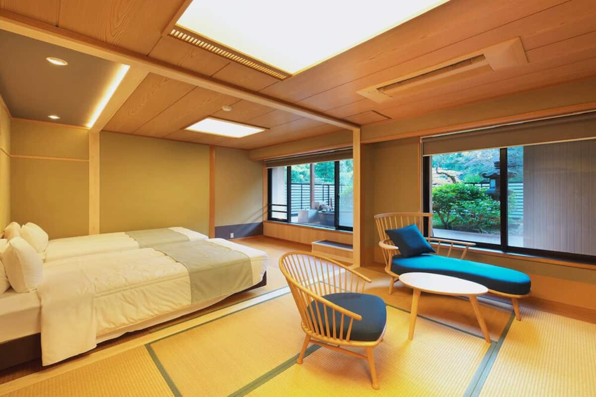 A room with a bed, a chair, a small table, and large windows.