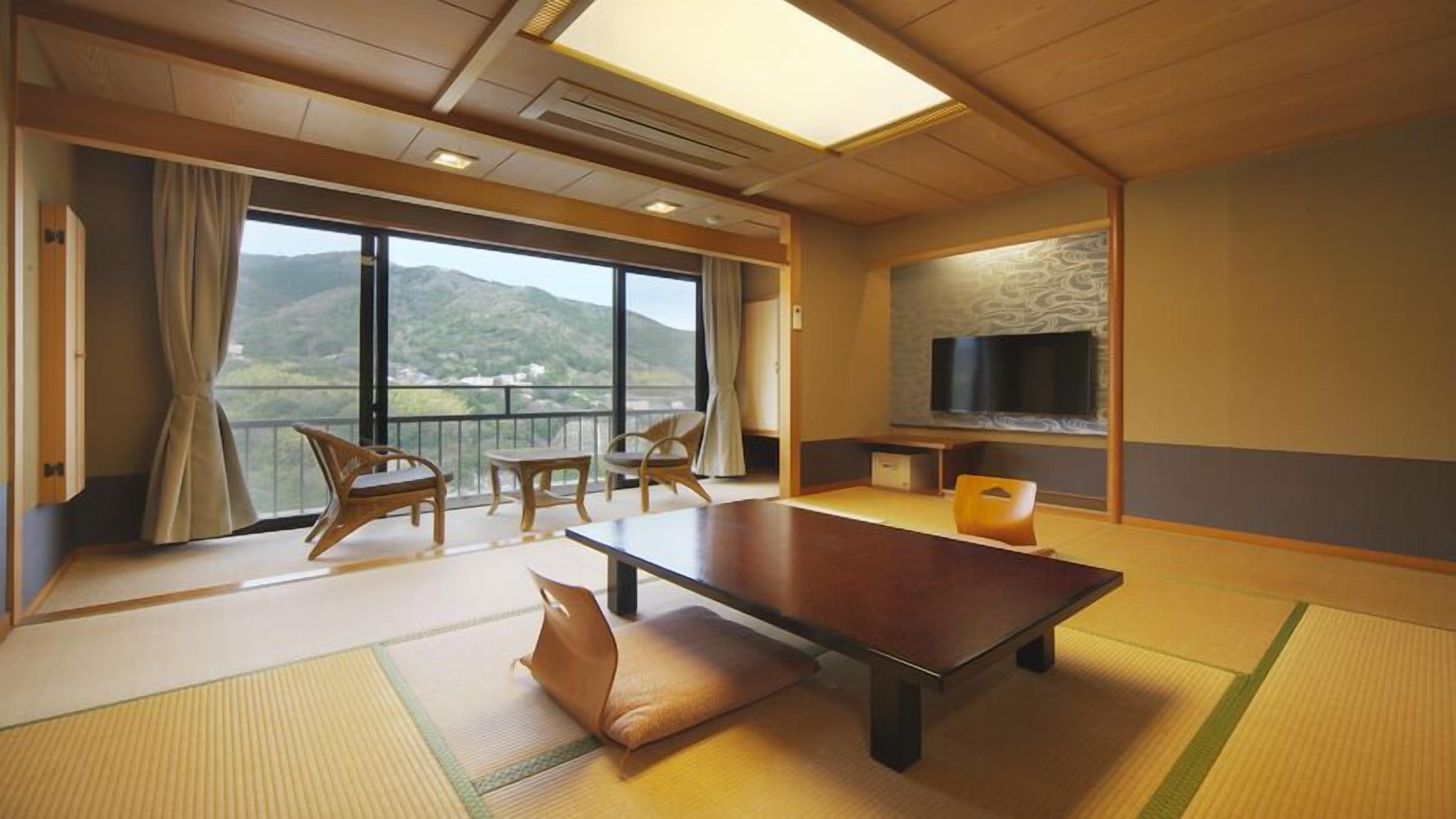 A room with tatami flooring, a low table, chairs, a sofa, a TV, and a balcony with a view of hills.