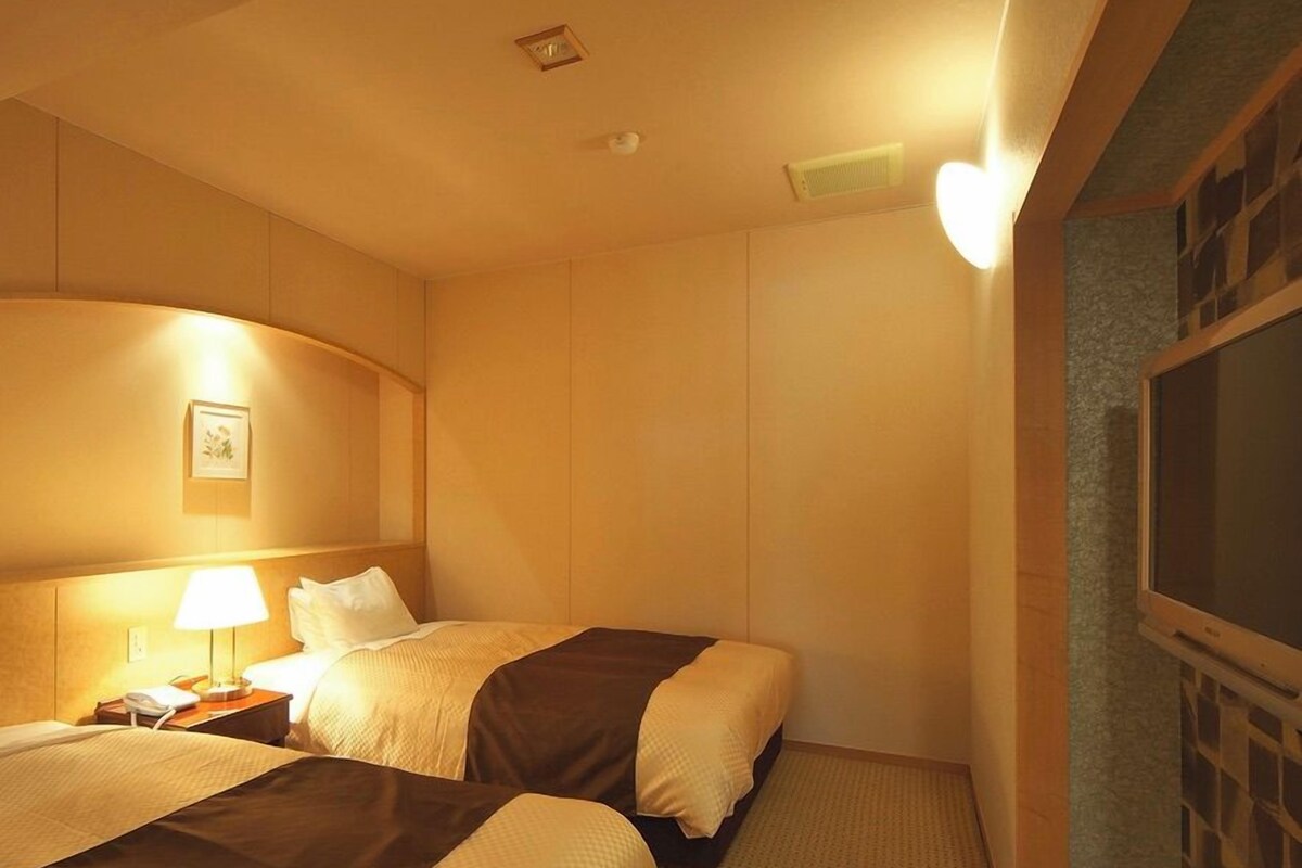 A hotel room with two beds, a nightstand with a lamp, a wall-mounted TV, and a framed picture.