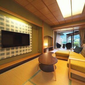 Habitación de hotel con televisor de pantalla plana en la pared, un sofá, una mesita redonda y vistas al exterior a través de amplias ventanas.