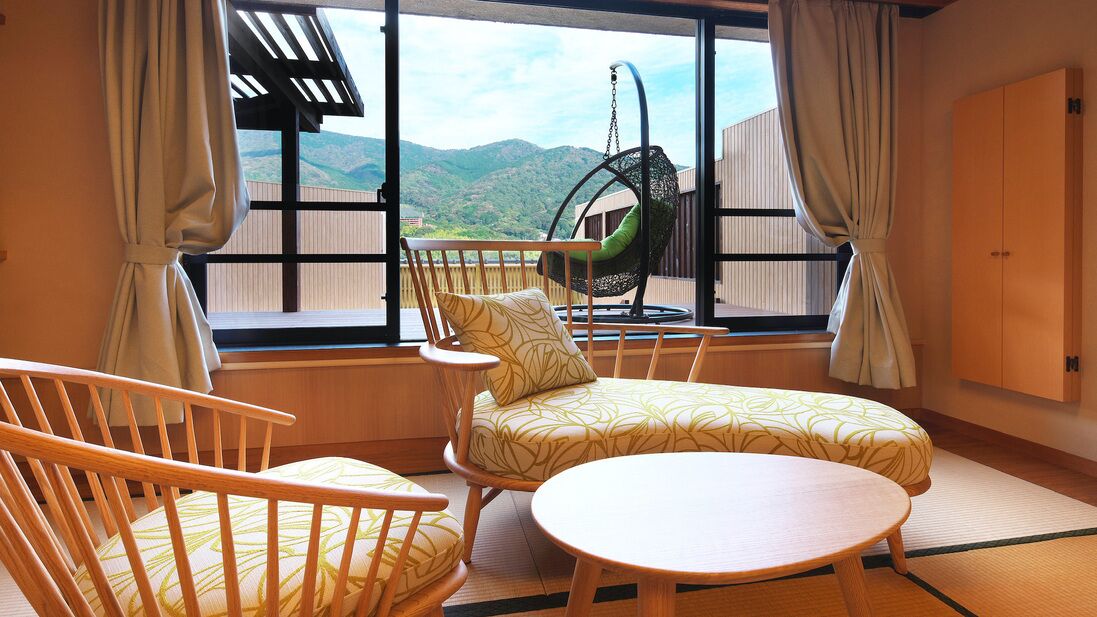A room with a large window, a hanging chair, a sofa, a round table, and a wooden chair.