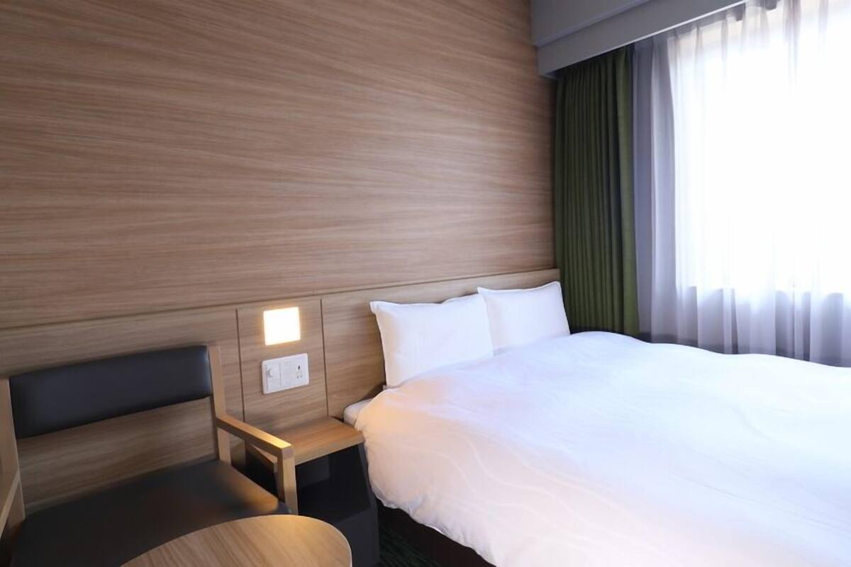 A hotel room with a wooden headboard, a bedside table with a lamp, a large bed, and a window with curtains.