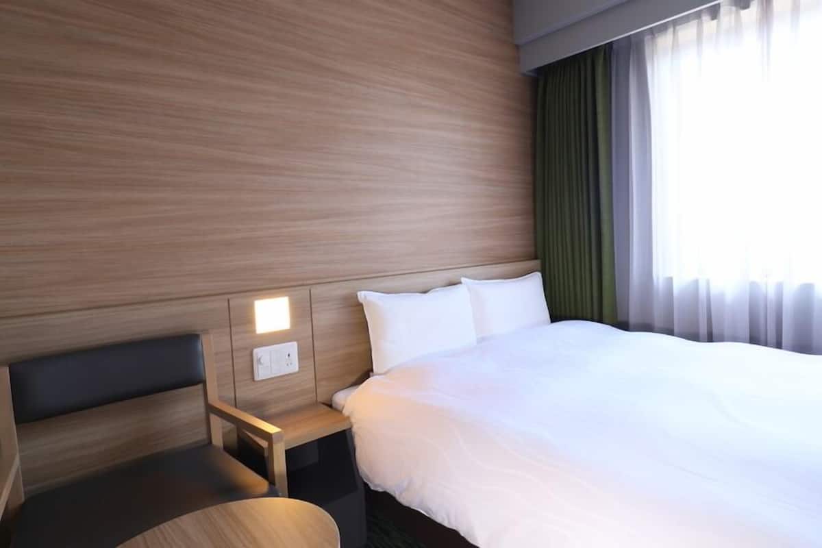 A hotel room with a wooden headboard, a bedside table with a lamp, a large bed, and a window with curtains.