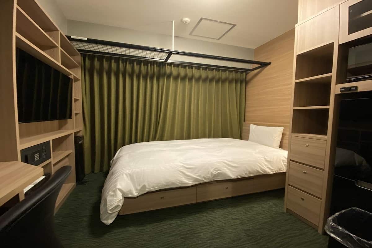 A bedroom with a bed, a desk, a wardrobe, and a trash bin.