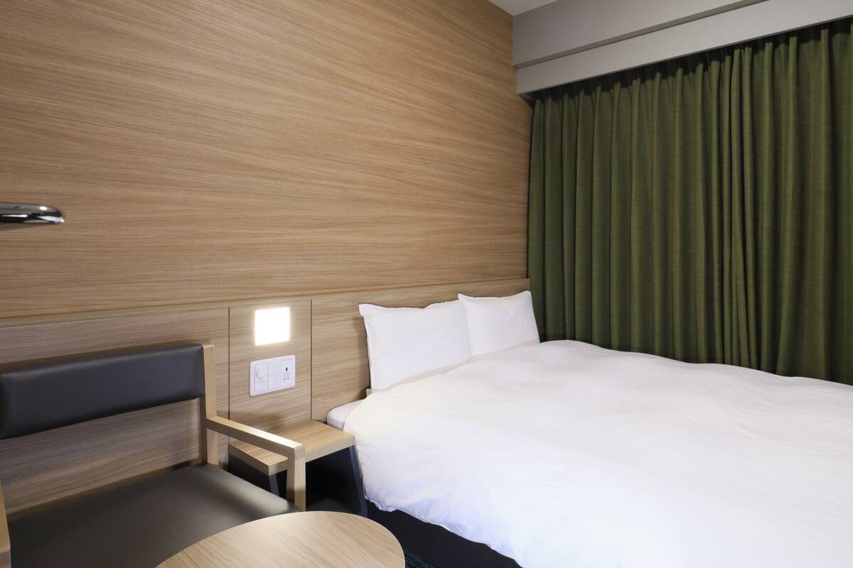 A hotel room with a bed, a nightstand, a lamp, a window with curtains, and a wooden wall panel.