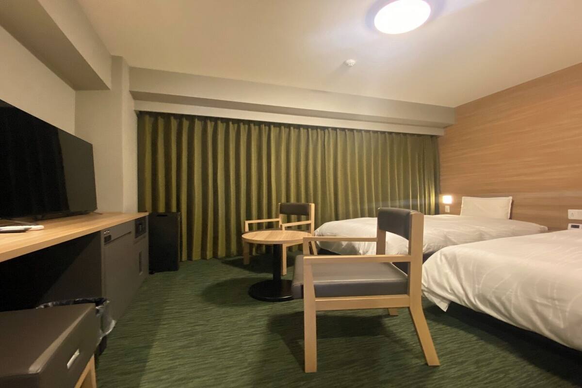 A hotel room with a TV, a desk, a chair, two beds, and a curtain.