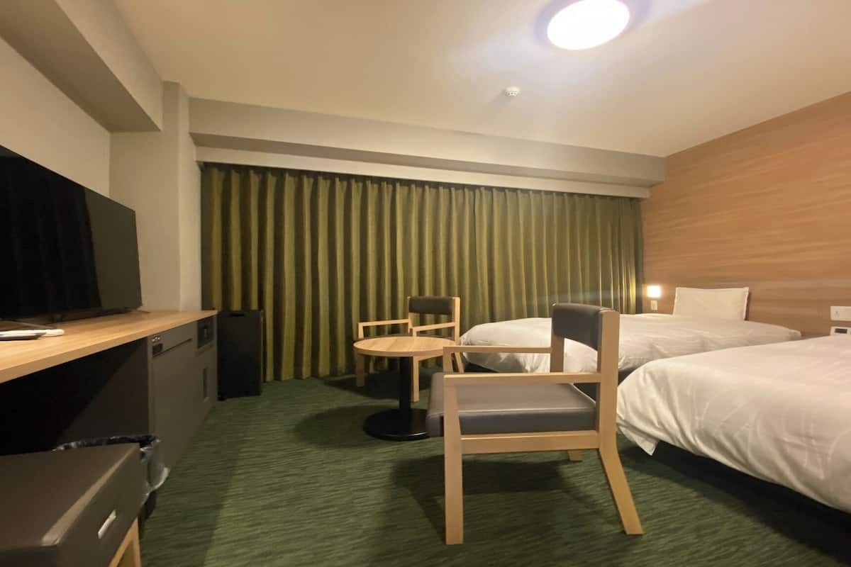 A hotel room with a TV, a desk, a chair, two beds, and a curtain.