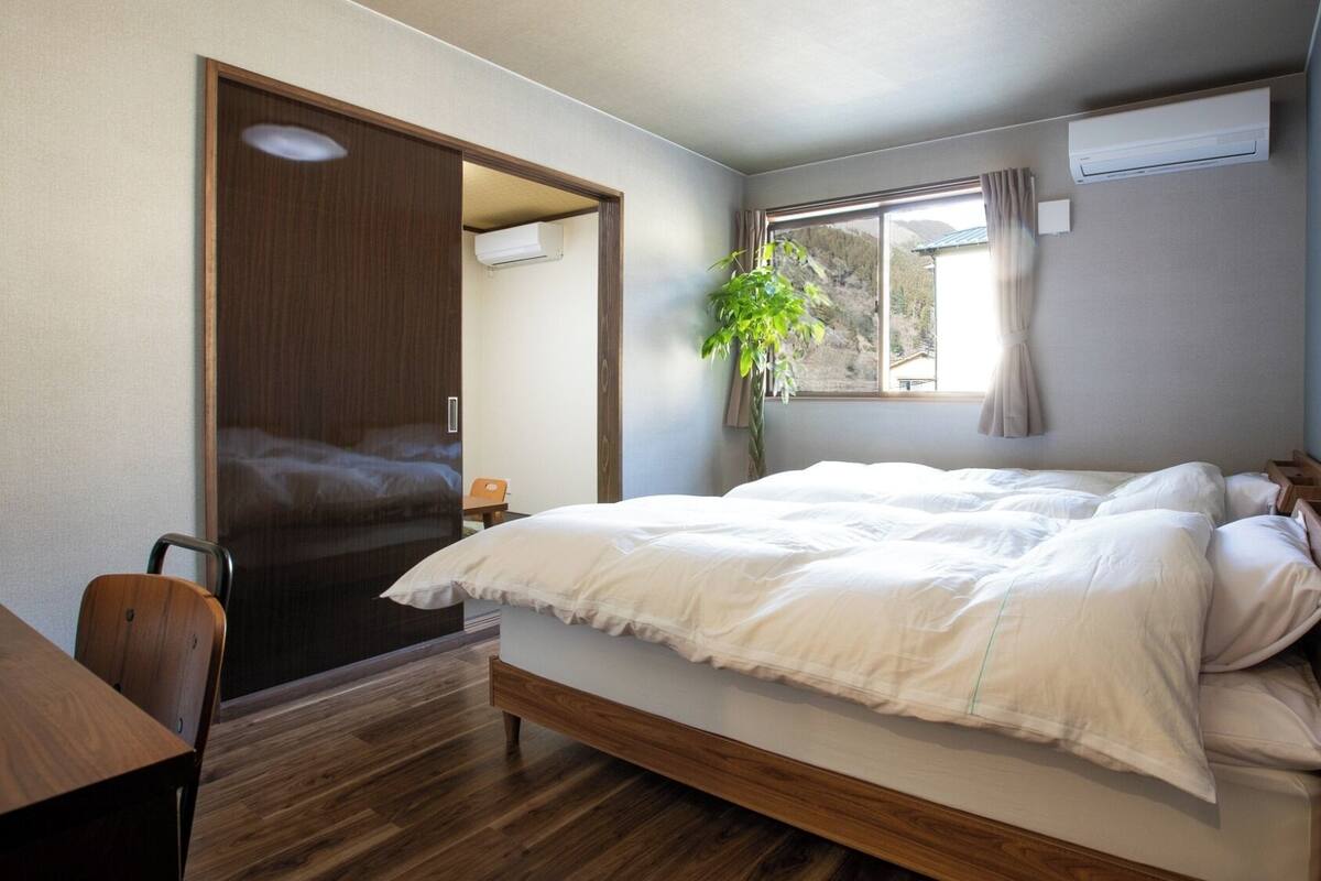 A bedroom with a bed, wooden floor, a desk, a chair, a plant, and an air conditioning unit.