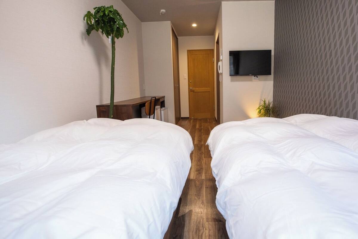 A hotel room with two beds, a desk, a chair, a potted plant, a TV, and a door leading to another room.
