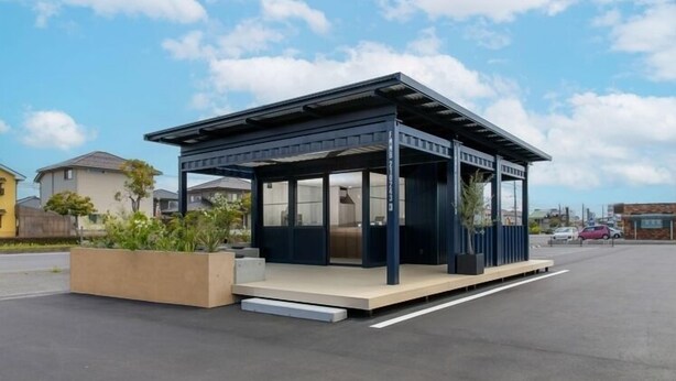 Une petite structure moderne de type pavillon, dotée d’un toit plat et de grandes fenêtres en verre, située sur un parking par beau temps.