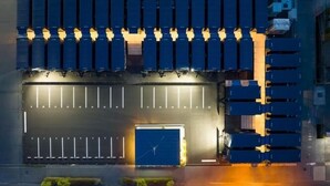 Une vue aérienne d’un parking où l’on aperçoit des rangées de conteneurs maritimes bleus, une grande porte de garage bleue au centre et une route dégagée devant.