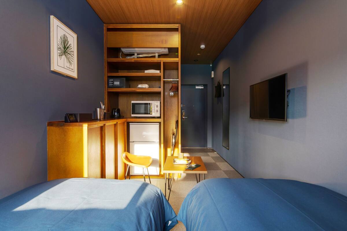 Habitación de hotel compacta con dos camas, un kitchenette, una pequeña mesa y un televisor montado en la pared.