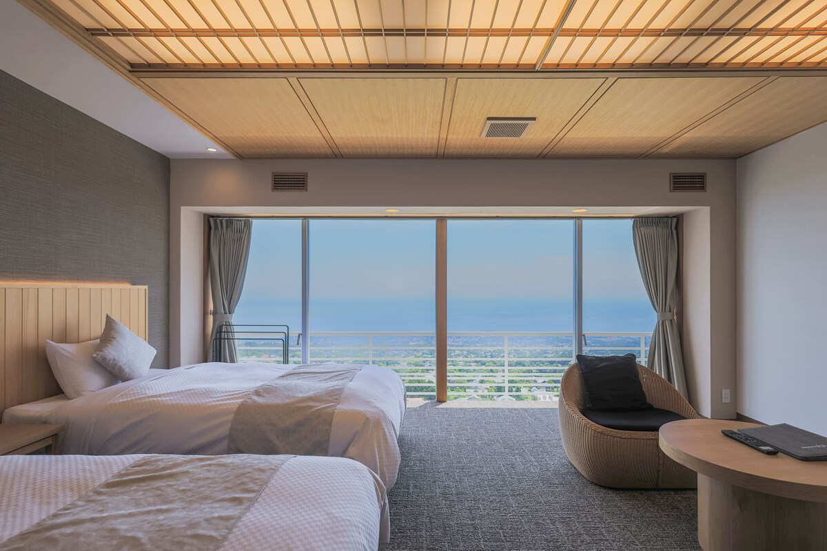 A hotel room with a large bed, a chair, a small table, and a view of the cityscape through large windows.