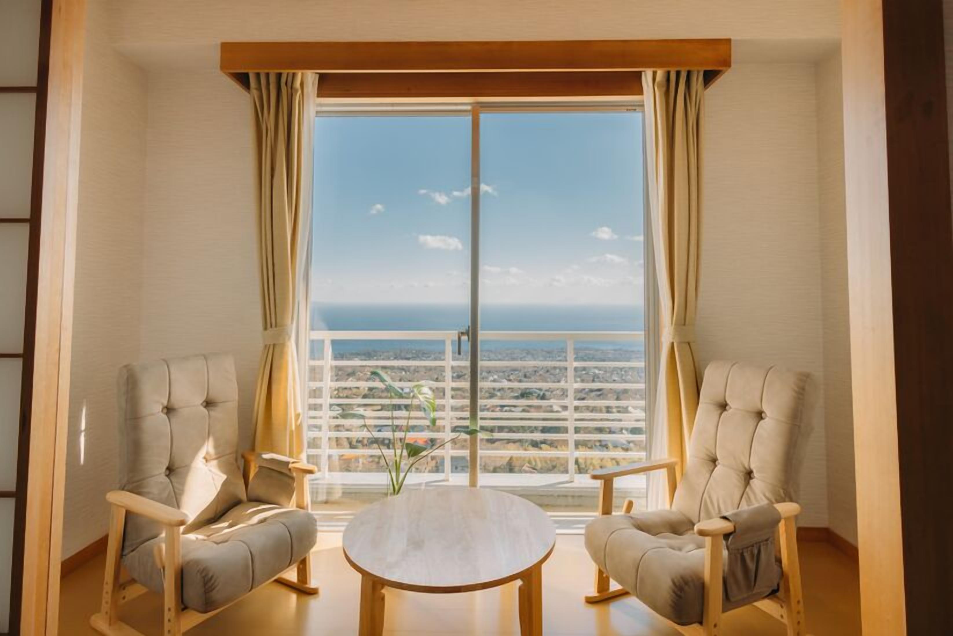 A room with two upholstered chairs, a round wooden table, and a large window with a view of a cityscape.