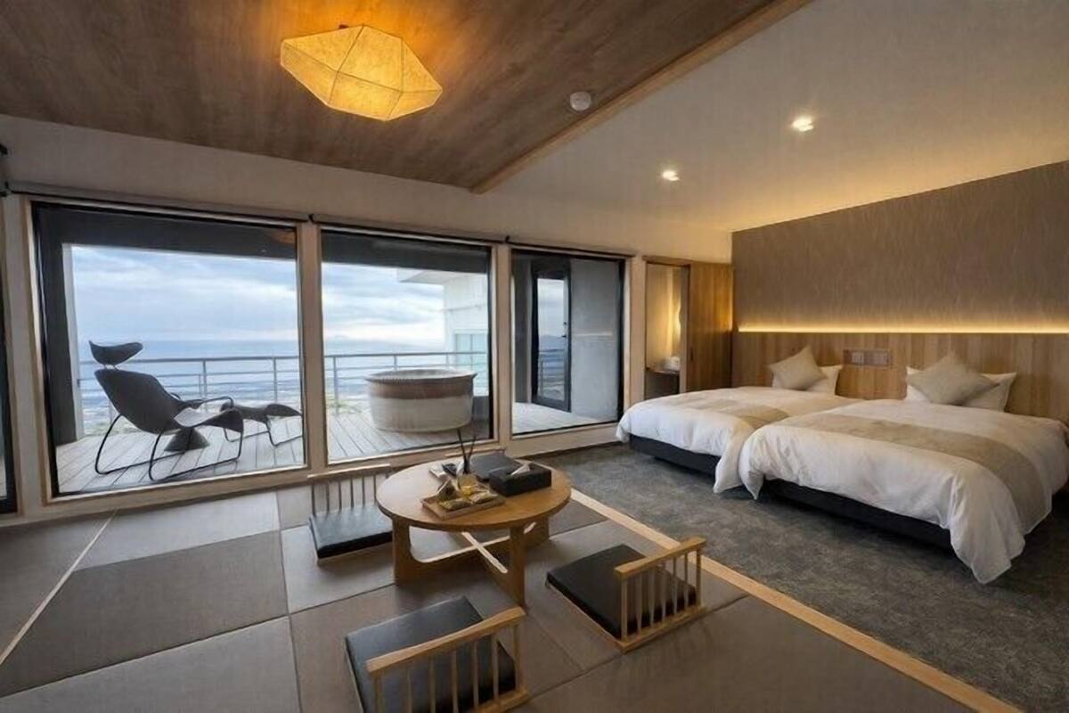 Une chambre d’hôtel moderne dotée d’un grand lit, d’un coin salon avec une table et d’une vue sur l’océan grâce à de grandes fenêtres.