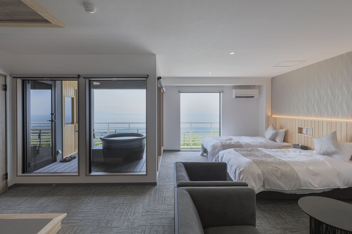 A modern hotel room with a balcony, a large bed, a seating area, and a view of the sea.