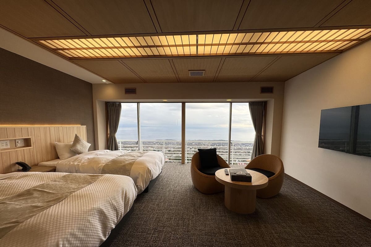 Une chambre d’hôtel avec deux lits, un canapé, une petite table et une grande fenêtre donnant sur la ville.