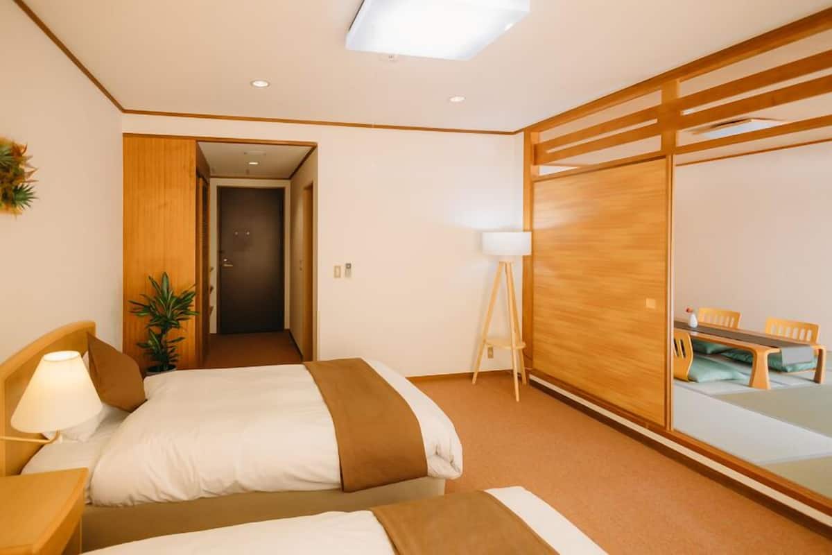 Une chambre d’hôtel avec deux lits, un bureau, une lampe, une plante et une porte coulissante donnant sur un coin repas.