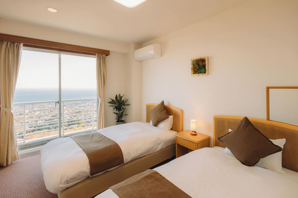 Une chambre d’hôtel avec deux lits, une grande fenêtre donnant sur la ville, un appareil de climatisation et une table de chevet avec une lampe.
